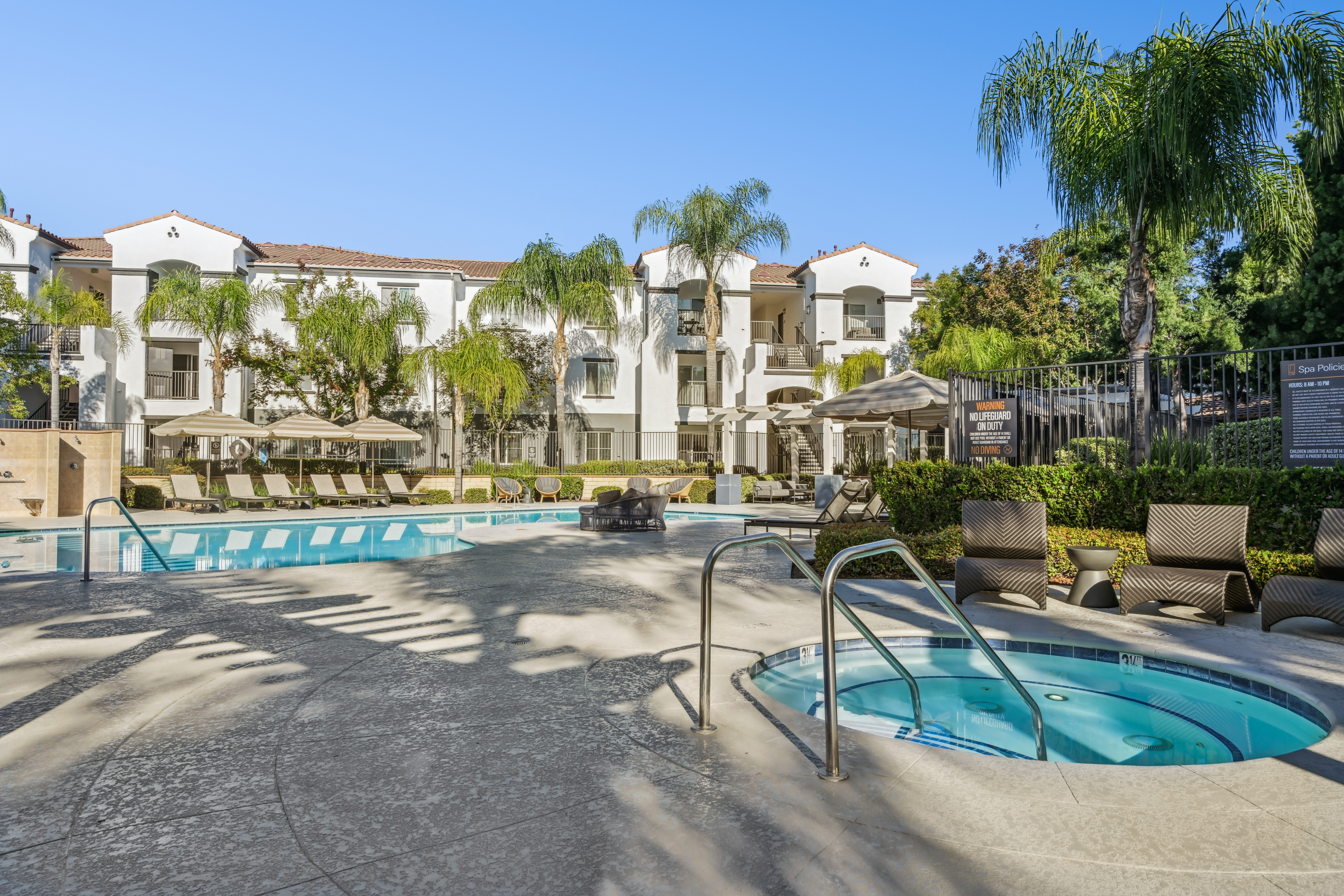 A sunny outdoor pool area with lounge chairs, umbrellas, a hot tub in the foreground, palm trees, and a white Mediterranean-style apartment building in the background.